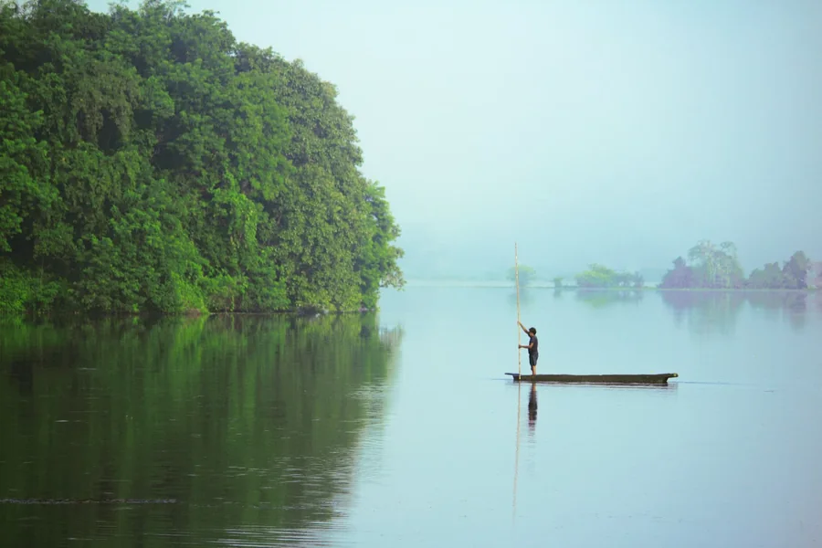 A fisherman crossing a still lake on a narrow wooden boat